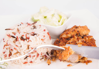 Pork chop (covered in breadcrumbs), three color rice and cucumber salad being eaten with a fork and a knife