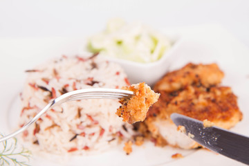 Pork chop (covered in breadcrumbs), three color rice and cucumber salad being eaten with a fork and a knife