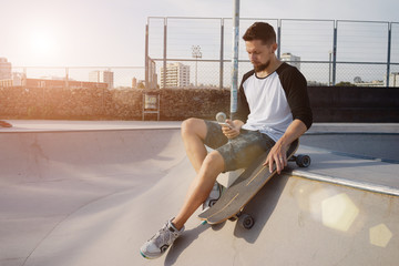 Young man is checking the email on his mobile phone while sitting in a skate park. A male with a skateboard is using the smartphone while having rest in a skating zone. Flare light. © JKstock