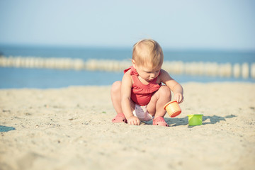 Baby playing on the sandy beach near the sea. Cute little girl in red dress with sand on tropical beach. Ocean coast.