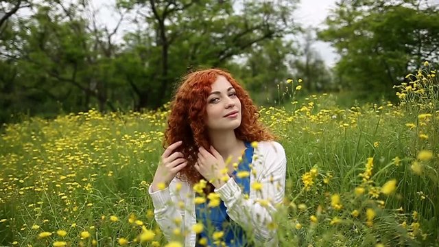 Cute red-haired girl straightens curly hair. Girl sits on a meadow among the wildflowers. Female face close up. Slow motion