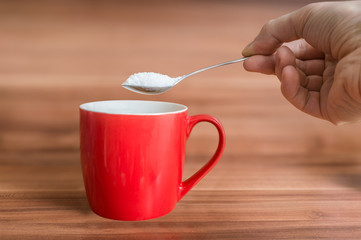 Hand holds teaspoon with sugar above red cup of tea. Unhealthy eating concept.