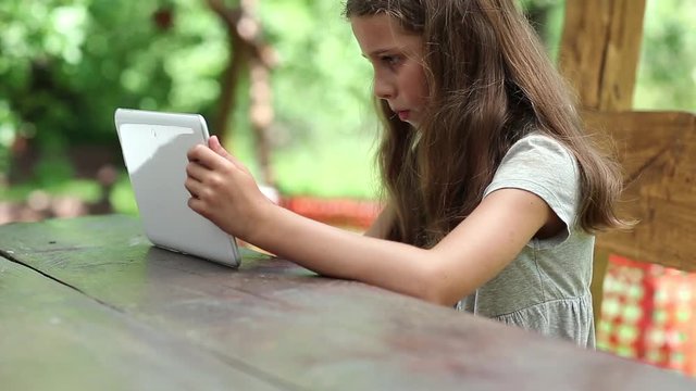 Beautiful girl with white tablet computer sits at the table. Girl plays the game on his Tablet PC. Little girl with tablet computer sits on the bench in garden. Female with tablet computer