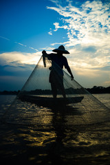 Silhouette of asian fisherman on wooden boat in action casting a net for catching freshwater fish...