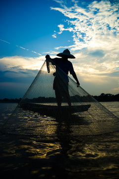 Silhouette of asian fisherman on wooden boat in action casting a net for catching freshwater fish in nature river in the early morning before sunrise