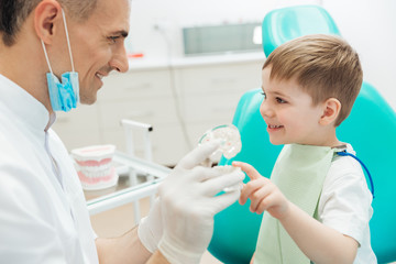 Cheerful man dentist demonstrating tooth brushing to little boy