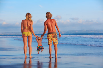 Happy family - father, mother, baby son hold hands, swim with fun, walk along sunset sea surf on black sand beach. Travel, active parents lifestyle, people activity on summer vacations with children.