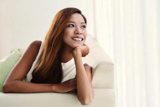 Smiling pensive Asian woman sitting on couch