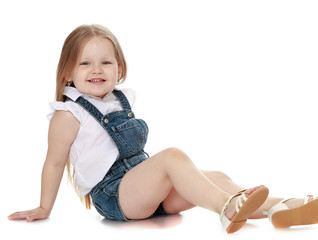 Laughing little girl with long blond hair in denim overalls sitting on the floor - Isolated on white background