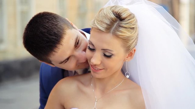 Close-up beautiful blonde bride smiling while groom whispering compliments to her and kissing