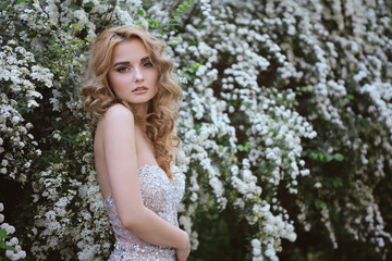 Fototapeta na wymiar young beautiful curly blonde hair slim girl fashion portrait in white dress posing looking into the camera calm look, in background lush bush with white flowers. Healthy woman lifestyle concept. 
