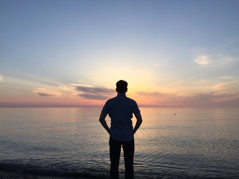 Young thoughtful man standing at the beach looking to the sunset or sunrise and dreaming about the future. Rear view.