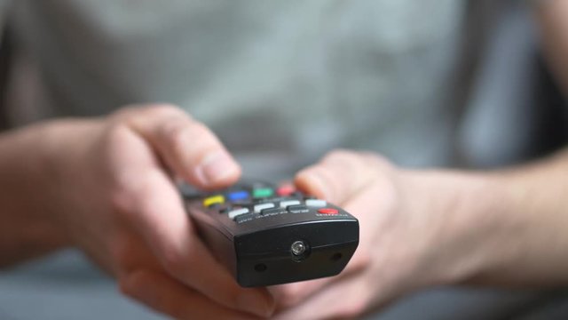 Male hand using a black casual tv remote controle to switch channels on television.