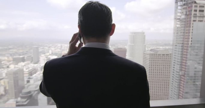 Successful young business man using a cellphone in front of a skyscraper window with views across Los Angeles.  Medium close up, rear view, dolly shot originally recorded in 4K