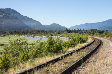 Fototapeta premium Rail tracks by a swampy lake surounded by mountains. Taken in Squamish, British Columbia, Canada on a sunny day.