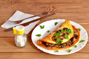 Omelet with mushrooms, cheese and parsley. Omelet with stuffing on a plate and on a wooden table. Healthy and tasty breakfast. Fork, knife, napkin, salt shaker.