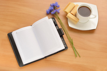 opened notebook with a cup of tea and flower on wooden table. Top view.