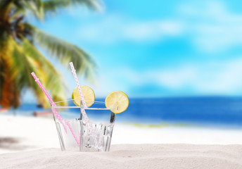 Cold water drinks with ice and a slice of lime on sand with tropical beach background