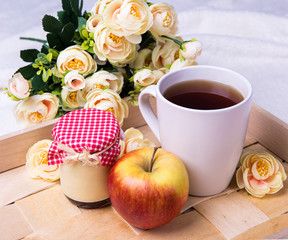wooden tray with cup of tea, jam-jar, apple and rose flowers