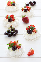 Homemade small pavlova meringue cakes with mascarpone cream, strawberries, cherries and fresh mint leaves on white wooden background