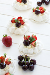 Homemade small pavlova meringue cakes with mascarpone cream, strawberries, cherries and fresh mint leaves on white wooden background