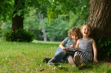Naklejka na ściany i meble Boy with girl of 7-8 years sit under a tree having nestled and play on the tablet.