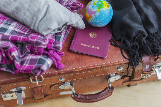 High Angle Close Up of Travel Vacation Concept Image - Brown Leather Suitcase with Plaid Pyjama Pants, Black Fringed Pashmina Wrap, Miniature Globe and German Passport