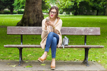 Cute young girl sitting on the bench and using smartphone in the park