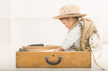Adorable little girl in a safari hat and explorer clothes reading old book sitting in a wooden suitcase with backpack and safety rope