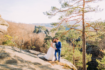 Handsome groom is gently holding his elegant new wife standing on rock slope