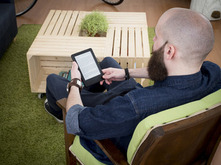 Young bearded man reading e-book at home. Shot from behind.