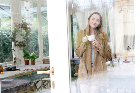 Woman in conservatory holding coffee cup looking away smiling