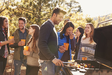 Group of friends stand at a barbecue, one cooking at grill - obrazy, fototapety, plakaty