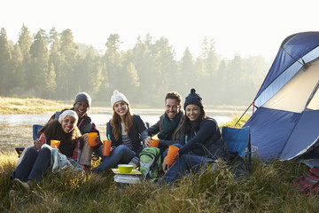 Friends sitting outside a tent near a lake looking to camera © Monkey Business
