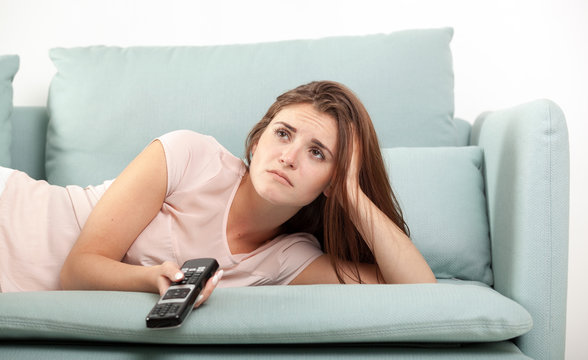 Sad young woman sitting on couch at home and watching TV. Casual style indoor shoot