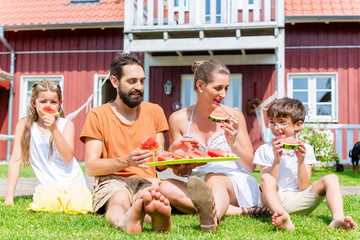 Familie sitzt vor Haus und isst Wassermelone im Garten