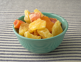 Bowl of sugared pineapple, mango and papaya on a blue striped tablecloth illuminated with window light.