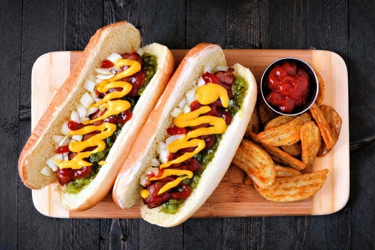 Two Hot Dogs Fully Loaded With Toppings And Potato Wedges On Wooden Board, Overhead View