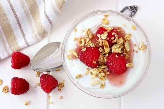 Raspberry flavored Greek yogurt with granola, overhead view with spoon on white wood