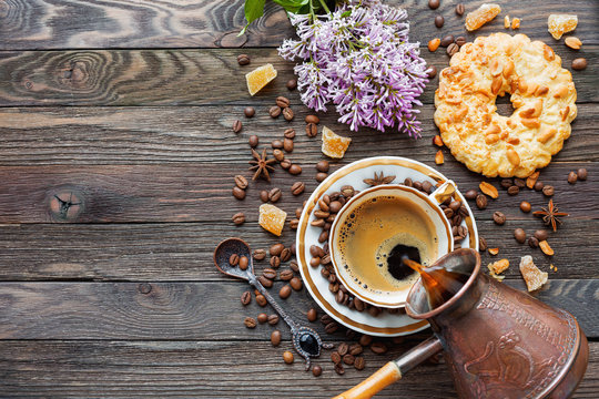 Rustic wooden background with cup and cezve of coffee, peanut tart and lilac flowers. White vintage dinnerware and spoon. Breakfast at summer morning. Top view, place for text.