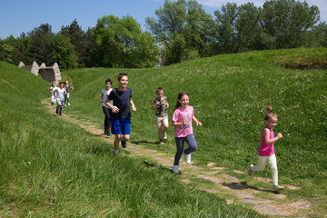 Obraz premium Happy And Smiling Group Of Kids Running In The Green Valley At Sunny Beautiful Day, Forest Trees And Blue Cloudy Sky In The Background