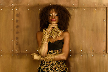Thoughtful young African woman in gold makeup and accessories holding hand on chin, looking at camera
