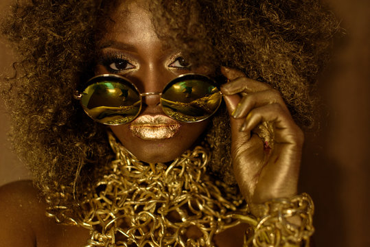 Beautiful African Or Black American Woman Wearing Gold Makeup And Accessories Looking Above Sunglasses, Studio Shot