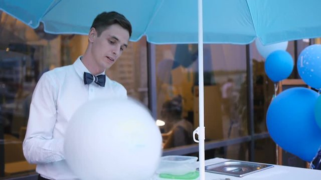 Young guy making cotton candy on a special machine, it wears bow tie, behind him balloons
