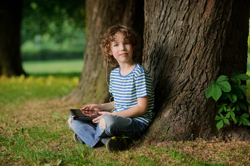 Boy of 8-9 years sits leaning against a tree and holds the tablet in hand.