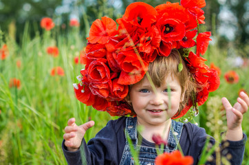 Little girl baby playing happy on the poppy field with a wreath, a bouquet of color A red poppies and white daisies, wearing a denim dress blue, charming, joyful child with a smile on his face