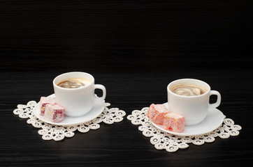 Side view of two mugs of coffee with ice-cream, Turkish delight on a saucer, on white lace napkins. Black background