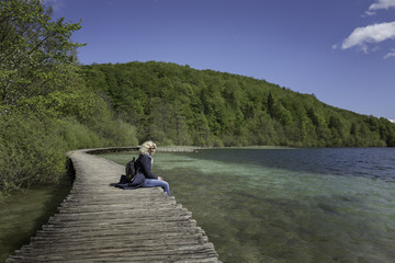 Blonde girl is seating on the bridge near of the lake with stunning blue water