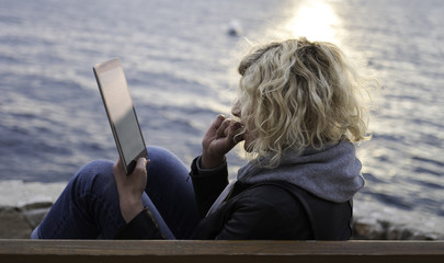 Blonde girl with curly hair is seating on the bench and absorbedly watching something in tablet