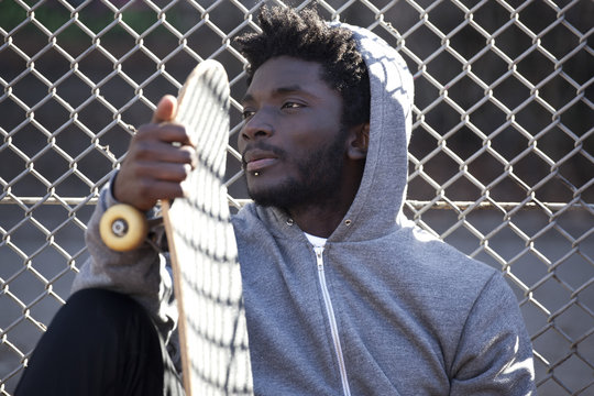 A serious young man leaning against a chain link fence holding a skateboard